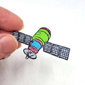May include: An embroidered patch depicting a colourful satellite with a green body, pink and blue accents, and two grey solar panels.