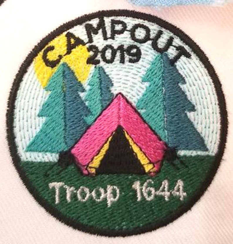 May include: A green and white embroidered patch with the text "Campout 2019" and "Troop 1644". The patch features a pink tent with a yellow and black interior, surrounded by three green trees and a yellow sun.