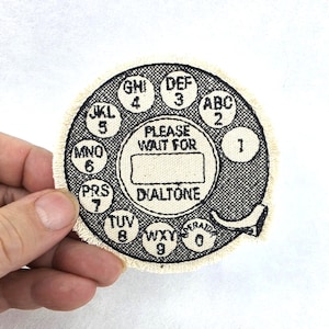 May include: A circular embroidered patch featuring a black rotary telephone dial design on a cream-colored fabric base. The patch includes black text reading "PLEASE WAIT FOR DIALTONE" in the center, surrounded by numbers 0-9 and letters.