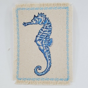 May include: A blue embroidered seahorse design on a beige fabric coaster with a blue and white border.