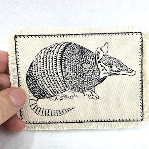 Puede incluir: Un parche rectangular de tela color hueso con una ilustración bordada en negro de un armadillo. El parche tiene bordes deshilachados y un marco con costura negra, resaltando la textura detallada del caparazón y la cola del animal.