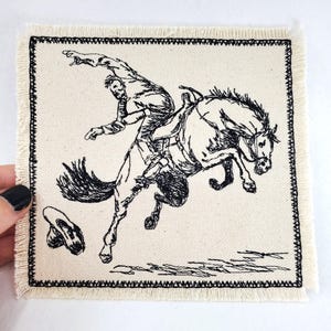 Könnte beinhalten: Schwarz-weiß gestickter Patch mit einem Cowboy, der ein sich aufbäumendes Pferd reitet. Das Pferd bäumt sich auf und der Cowboy hält sich mit einer Hand fest. Der Patch hat einen ausgefransten Rand.