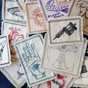 May include: A collection of embroidered patches featuring various designs, including a hand with an eye, a heart with thorns, a brain, a sun, a fish, a tattoo machine, a Vitruvian Man, and a bee. The patches are made of beige fabric with frayed edges and are decorated with colorful embroidery.
