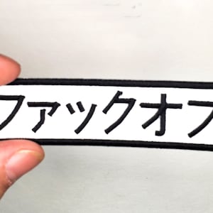 May include: A white rectangular patch with a black border and the Japanese text "ファックオフ" embroidered in black.