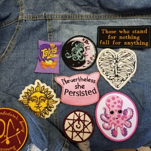 May include: A collection of embroidered patches on a blue denim jacket. Patches include a Takis bag, a moon face, a ribcage heart, a sun face, a pink cat-ear shape with text 'Nevertheless she Persisted', a symbol, an octopus, and a Dumbledore's Army logo.