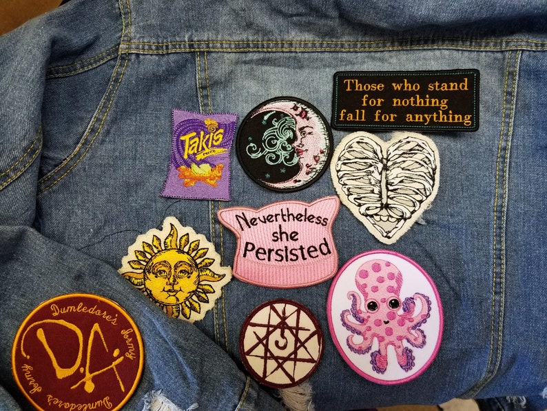 May include: A collection of embroidered patches on a blue denim jacket. Patches include a Takis bag, a moon face, a ribcage heart, a sun face, a pink cat-ear shape with text 'Nevertheless she Persisted', a symbol, an octopus, and a Dumbledore's Army logo.