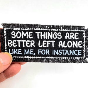 May include: Black rectangular patch with white embroidered text: "SOME THINGS ARE BETTER LEFT ALONE LIKE ME, FOR INSTANCE." The patch has a white border and a frayed edge.