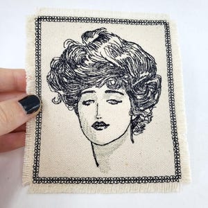 Puede incluir: Un parche bordado en blanco y negro que representa a una mujer con un peinado grande y elaborado. El parche está enmarcado con un borde cosido negro y tiene un borde deshilachado.