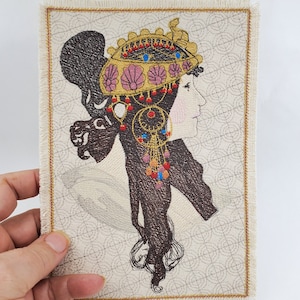 Peut inclure: Patch brodé représentant une femme aux longs cheveux noirs portant une coiffe dorée et rose avec des accents rouges et bleus. L'arrière-plan est un tissu crème avec un motif répétitif.