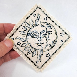 Peut inclure: Écusson brodé en forme de losange avec un motif de soleil et de lune avec un visage, de couleur bleu marine sur fond crème. Le patch est orné d'étoiles et a un bord effiloché. Thème céleste.