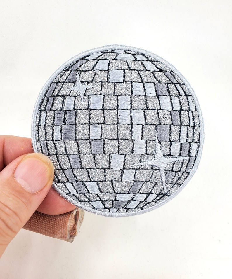 Puede incluir: Parche bordado con un dise&ntilde;o de bola de discoteca plateada, con un patr&oacute;n de cuadr&iacute;cula y dos acentos de estrella. El parche tiene un borde gris y lo sostiene una mano sobre un fondo blanco. El parche es redondo.