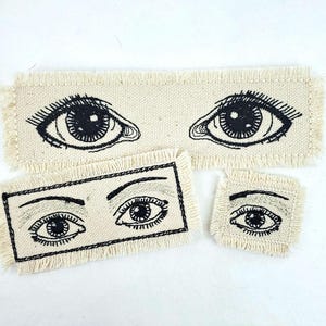 Pode incluir: Três patches de tecido bordados com olhos pretos sobre um fundo branco. Os patches são de tamanhos diferentes e têm bordas desfiadas.