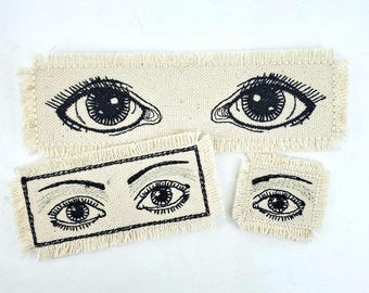 May include: Three embroidered fabric patches with black eyes on a white background. The patches are different sizes and have frayed edges.