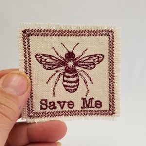 May include: A beige fabric patch with a red embroidered border. The patch features a red embroidered bee with the text "Save Me" below it.