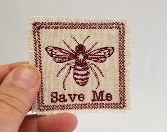 May include: A beige fabric patch with a red embroidered border. The patch features a red embroidered bee with the text "Save Me" below it.