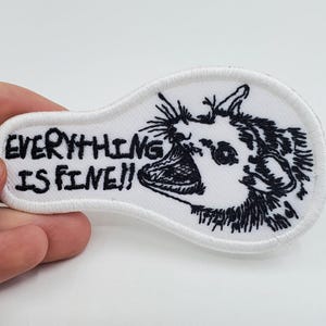 Puede incluir: Parche blanco bordado con puntadas negras. El parche presenta un dibujo de estilo de dibujos animados de una criatura con la boca muy abierta y el texto "EVERYTHING IS FINE!"