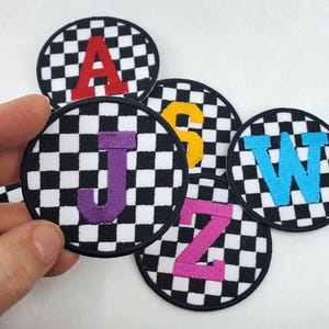 Peut inclure: Écussons brodés ronds avec un motif à carreaux noir et blanc et des lettres colorées. Les lettres comprennent A rouge, S jaune, J violet, W bleu et Z rose. Chaque écusson a une bordure noire.