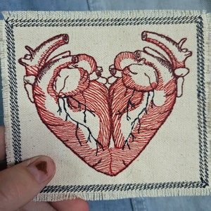 Puede incluir: Un parche bordado rojo que representa dos corazones anatómicos entrelazados en forma de corazón. El parche está cosido sobre una tela beige con un borde negro cosido.