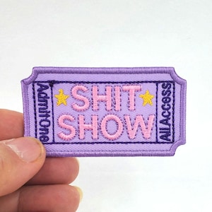 May include: Purple fabric patch with the text "Shit Show" in pink letters and "All Access" and "Admit One" on the sides. There are two yellow stars on either side of the text.