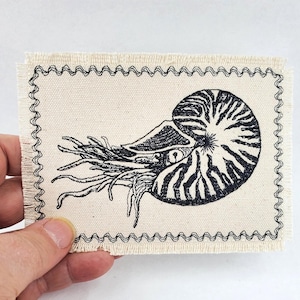 May include: A rectangular patch with a black and white nautilus illustration. The patch has a cream-colored background with a black zigzag stitched border. The nautilus is detailed with black lines.