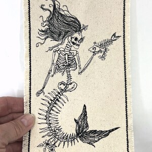May include: Black and white embroidered patch of a skeleton mermaid holding a fish skeleton. The mermaid has long flowing hair and a tail with floral details.