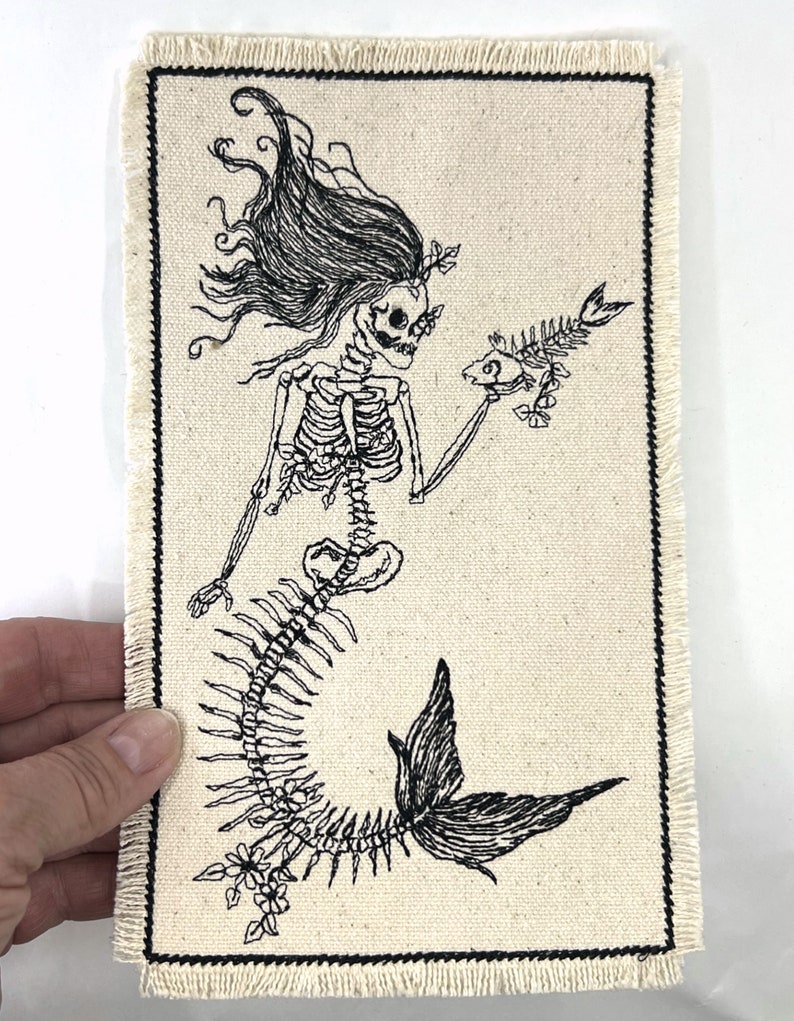 May include: Black and white embroidered patch of a skeleton mermaid holding a fish skeleton. The mermaid has long flowing hair and a tail with floral details.