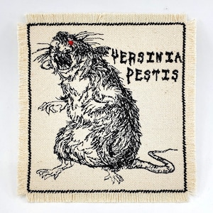 Puede incluir: Un parche bordado en blanco y negro con una rata y el texto "Yersinia Pestis". La rata tiene un ojo rojo.