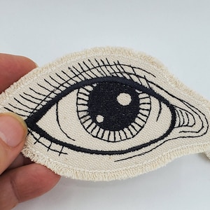 May include: Two embroidered fabric patches with black thread depicting a large eye with eyelashes. The eyes are on a white background.