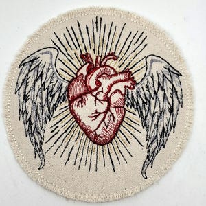 May include: Embroidered patch with a red anatomical heart with wings, surrounded by a golden halo of light rays on a beige background.