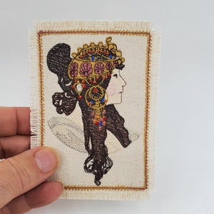 Peut inclure: Écusson brodé représentant le profil d'une femme aux cheveux foncés et coiffée d'un ornement de tête décoratif. L'ornement est doré avec des détails roses et bleus. Le patch a un fond crème, une bordure dorée et des bords effilochés.