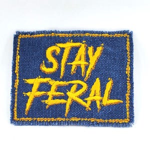 May include: A blue denim patch with yellow embroidered text that reads "Stay Feral".