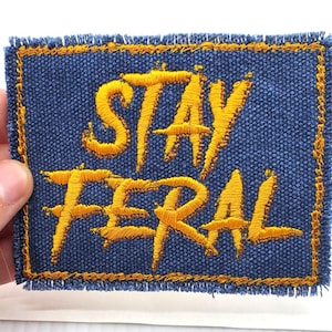 Puede incluir: Un parche de mezclilla azul con texto bordado en amarillo que dice "Stay Feral".