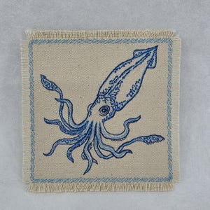 May include: A square beige fabric coaster with frayed edges featuring a blue embroidered squid design in the center. The coaster is framed by a thin blue embroidered border that follows the square shape, highlighting the detailed marine creature.