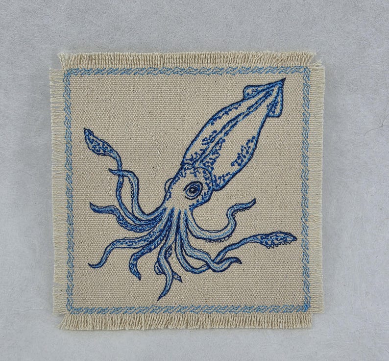 May include: A square beige fabric coaster with frayed edges featuring a blue embroidered squid design in the center. The coaster is framed by a thin blue embroidered border that follows the square shape, highlighting the detailed marine creature.