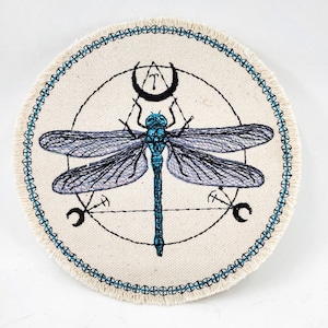May include: A round, white canvas patch with a blue dragonfly embroidered in the center. The dragonfly is surrounded by a geometric design with a crescent moon at the top.
