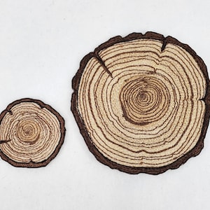 May include: Two wood slice coasters. The coasters feature a detailed design of tree rings in shades of brown and tan, with a dark brown border. One coaster is smaller than the other.