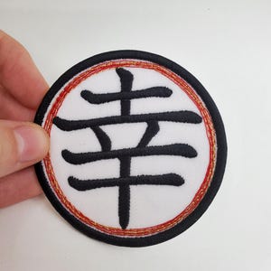 Puede incluir: Un parche bordado blanco con un carácter kanji negro en el centro. El carácter kanji está rodeado por un borde bordado rojo y dorado.