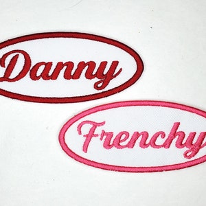May include: Two white oval embroidered patches with red and pink stitching. The top patch says "Danny" in red lettering. The bottom patch says "Frenchie" in pink lettering.