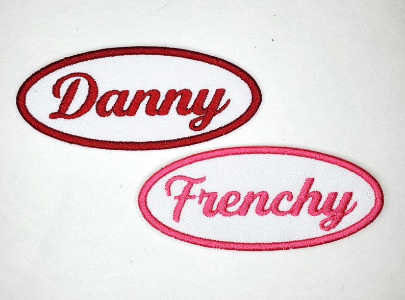 May include: Two white oval embroidered patches with red and pink stitching. The top patch says "Danny" in red lettering. The bottom patch says "Frenchie" in pink lettering.