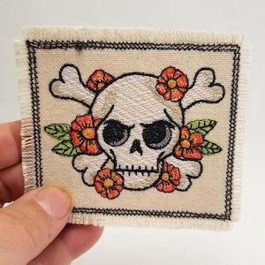May include: A square fabric patch with a frayed edge featuring an embroidered design of a white skull and crossbones surrounded by orange flowers and green leaves. The entire motif is framed by a black stitched border on a cream-colored background.