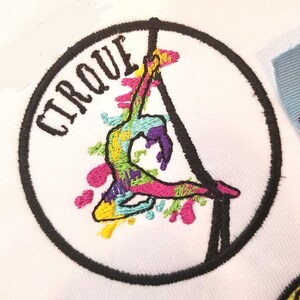 May include: A colorful embroidered patch with a circus performer hanging from a trapeze. The patch is circular and has a black border. The text "CIRQUE" is embroidered in black on the patch.