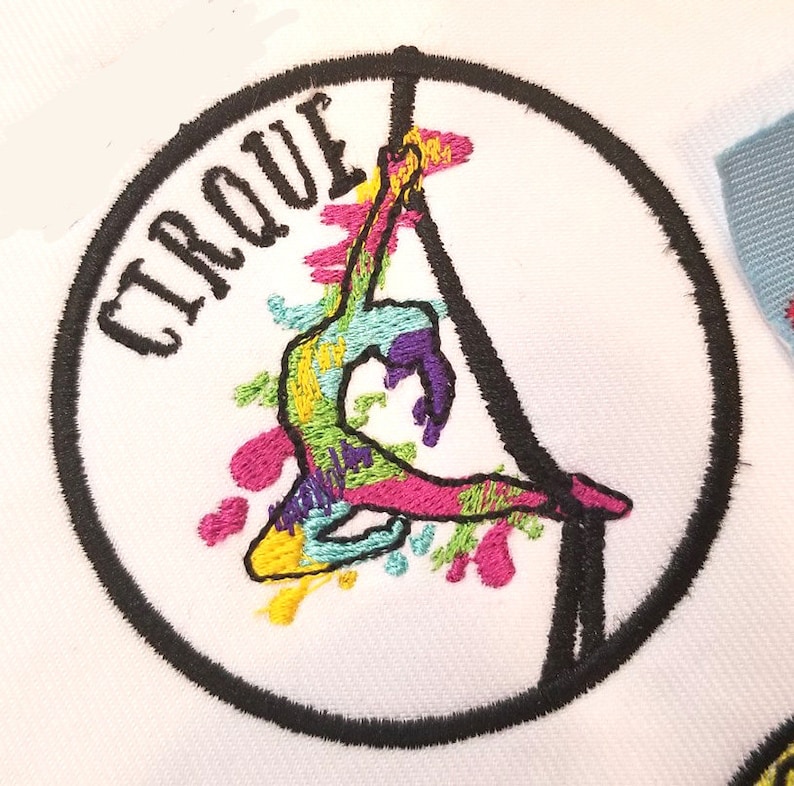 May include: A colorful embroidered patch with a circus performer hanging from a trapeze. The patch is circular and has a black border. The text "CIRQUE" is embroidered in black on the patch.