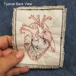 May include: A white fabric patch with a red embroidered anatomical heart design. The patch has a frayed edge and is surrounded by a black and white stitched border.