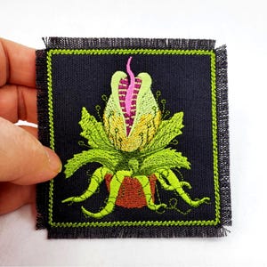 Puede incluir: Parche bordado que representa una planta estilizada con interior rosa y amarillo, hojas verdes y zarcillos. La planta está en una maceta marrón, sobre un fondo negro con un borde verde. El parche tiene un borde deshilachado.