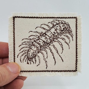 Könnte beinhalten: Ein brauner gestickter Aufnäher mit einem detaillierten Bild eines Tausendfüßlers auf einem weißen Stoffgrund. Der Aufnäher hat einen ausgefransten Rand.