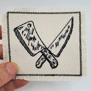 May include: A black and white embroidered patch featuring two crossed knives on a white canvas background. The patch has a frayed edge.