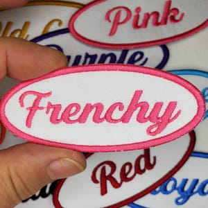 以下が含まれることがあります： ピンクの刺繍の縁取りが施された白い楕円形の布製パッチで、ピンクの筆記体で「Frenchie」と書いてあります。