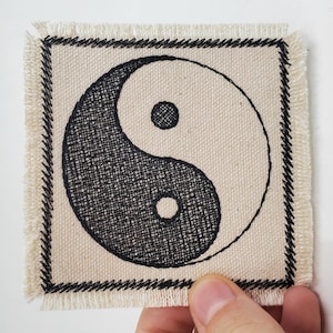 Pode incluir: Um remendo de tecido bege com um símbolo yin yang preto e branco bordado e uma borda preta costurada.
