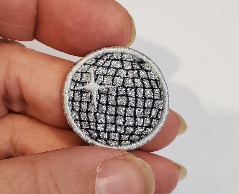 May include: A silver and black round embroidered patch with a geometric design. The patch has a white outline and a white star in the center.