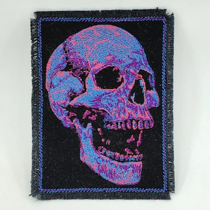May include: A black fabric patch with a pink and blue embroidered skull design. The skull is facing right and has an open mouth.